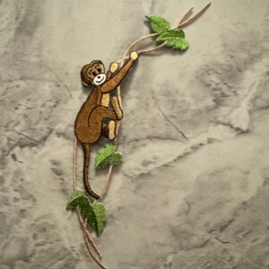 May include: Embroidered patch featuring a brown monkey clinging to a vine with green leaves. The monkey has a smiling face and is holding onto a thin, brown vine. The leaves are a bright green color.