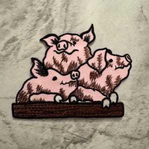 May include: Embroidered patch featuring two pink pigs, one on top of the other, with brown details. The pigs are sitting on a brown wooden log.