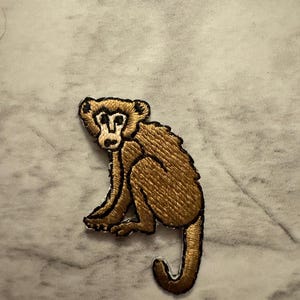 May include: Embroidered patch featuring a seated monkey with a brown body and a black outline. The monkey has a detailed design with visible fur texture. The patch is likely for clothing or accessories.
