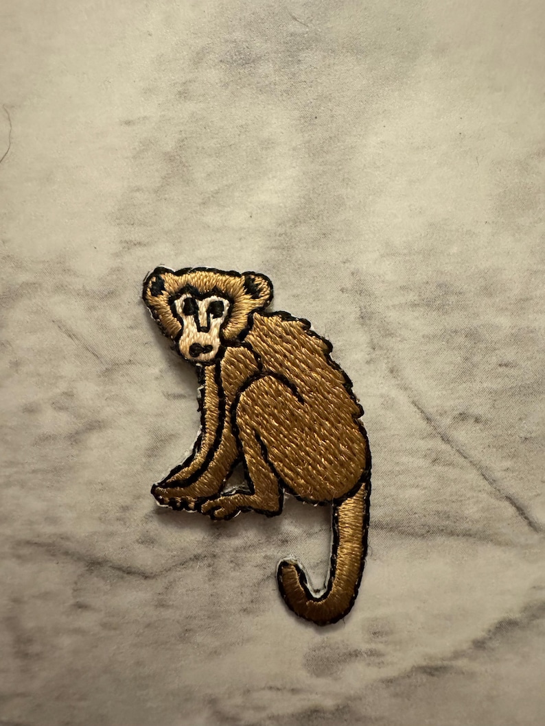 May include: Embroidered patch featuring a seated monkey with a brown body and a black outline. The monkey has a detailed design with visible fur texture. The patch is likely for clothing or accessories.