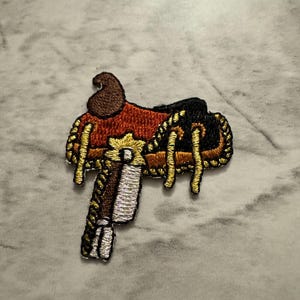 May include: Embroidered patch of a Western saddle. The patch features a brown, red, black, and gold color scheme. The saddle has intricate details, including gold rope-like trim and a detailed stirrup. The patch is likely designed for clothing or accessories.