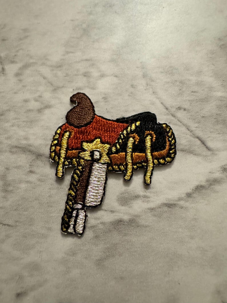 May include: Embroidered patch of a Western saddle. The patch features a brown, red, black, and gold color scheme. The saddle has intricate details, including gold rope-like trim and a detailed stirrup. The patch is likely designed for clothing or accessories.