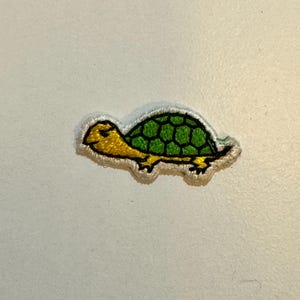 May include: A green and yellow embroidered turtle patch. The turtle is walking to the right and has a green shell with yellow accents.