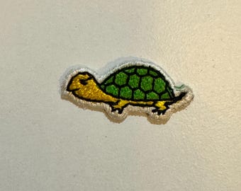 May include: A green and yellow embroidered turtle patch. The turtle is walking to the right and has a green shell with yellow accents.