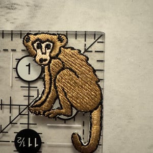 May include: Embroidered patch featuring a seated monkey in shades of brown and tan, with black outlining. The patch is on a clear ruler, showing measurements in inches. The monkey has a detailed face and a curled tail.