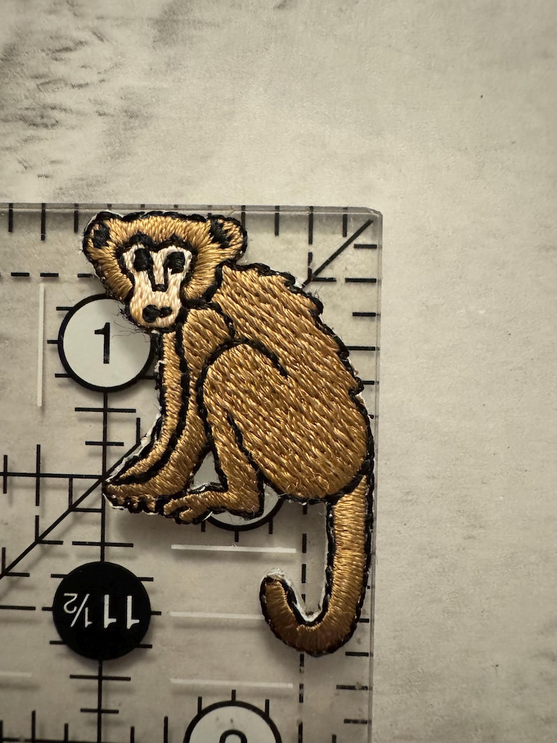 May include: Embroidered patch featuring a seated monkey in shades of brown and tan, with black outlining. The patch is on a clear ruler, showing measurements in inches. The monkey has a detailed face and a curled tail.