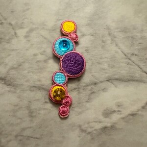 K&ouml;nnte beinhalten: Gestickter Aufn&auml;her mit einer Reihe farbiger Kreise in Gelb, T&uuml;rkis, Lila und Rosa. Jeder Kreis ist mit Pailletten oder Perlen verziert. Der Aufn&auml;her hat eine geschwungene, organische Form.