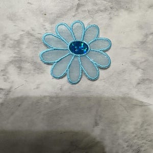 May include: A light blue flower-shaped patch featuring sheer, translucent petals with embroidered edges and a central oval embellishment filled with dark blue glitter. The design consists of twelve rounded petals arranged in a circular pattern.