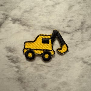 May include: A yellow embroidered patch of a construction excavator with black detailing. The excavator has a black arm and wheels, and a black window. The patch is on a gray and white marbled background.