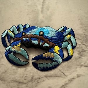 May include: Embroidered crab patch in shades of blue, turquoise, brown, and yellow. The crab has detailed stitching, with a textured appearance. The patch is likely for clothing or accessories.