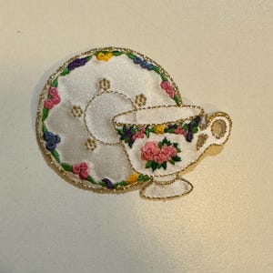 May include: A white embroidered teacup and saucer with pink, yellow, green, and purple flowers. The saucer has a gold trim.