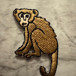 May include: Embroidered patch featuring a seated monkey. The monkey is rendered in shades of brown and gold, with black outlining the details. The patch is likely for clothing or accessories.