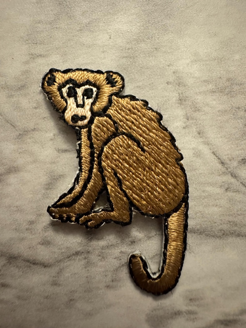 May include: Embroidered patch featuring a seated monkey. The monkey is rendered in shades of brown and gold, with black outlining the details. The patch is likely for clothing or accessories.