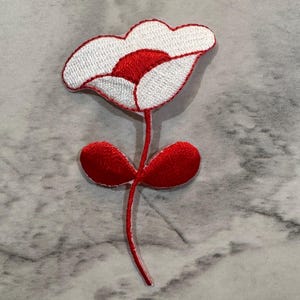 May include: Embroidered flower patch featuring a white flower with red accents. The flower has a red stem and two red leaves. The patch is designed to be sewn or ironed onto fabric. The flower patch is a decorative item.
