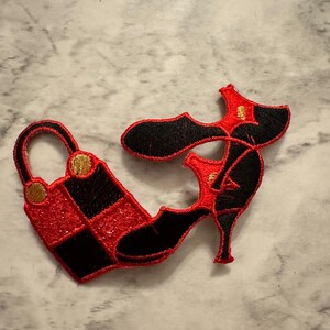 May include: Embroidered patch featuring a high-heeled shoe and a handbag. The shoe is black and red with a stiletto heel. The handbag is red with a black checkered pattern. Both patches have gold accents and are on a marble surface.
