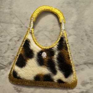 May include: A miniature handbag patch with a gold handle and trim. The bag features a cowhide pattern in black, brown, and white. The patch is embroidered with a gold thread.
