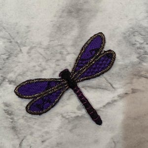 May include: A purple dragonfly patch with gold trim. The dragonfly has a black body and purple wings with a delicate lace-like pattern. The patch is likely for clothing or crafts.