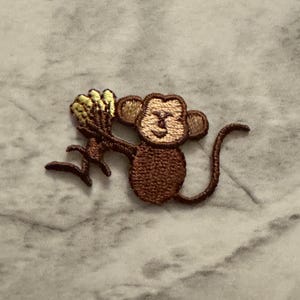 May include: Embroidered patch featuring a brown monkey holding a bunch of yellow bananas. The monkey has a smiling face and a long tail. The patch is designed for application on clothing or accessories.