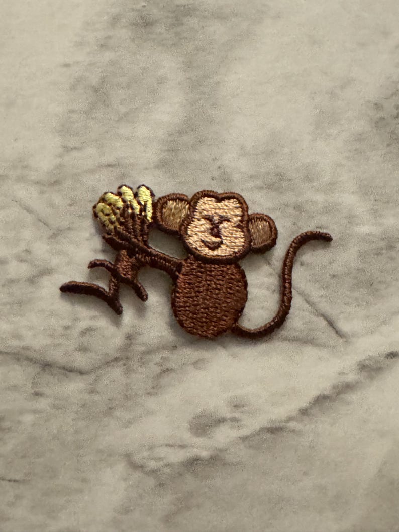 May include: Embroidered patch featuring a brown monkey holding a bunch of yellow bananas. The monkey has a smiling face and a long tail. The patch is designed for application on clothing or accessories.