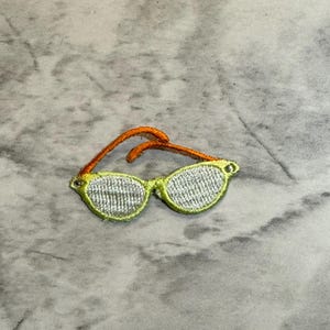May include: Embroidered patch of sunglasses with yellow frames and orange arms. The lenses are filled with a white, textured pattern. This patch is suitable for clothing, bags, or other fabric items.