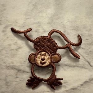 May include: Embroidered patch featuring a brown monkey with a light brown face and ears. The monkey is depicted with its arms and tail extended, giving it a playful appearance. The patch is likely designed to be ironed or sewn onto fabric.