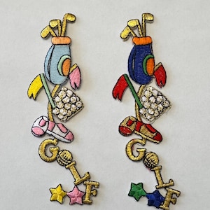 May include: Two embroidered iron-on patches with a golf theme. The patches are gold with colorful accents. The left patch has a light blue golf bag, pink shoe, yellow flag, and white golf balls. The right patch has a blue golf bag, red shoe, red flag, and white golf balls. Both patches have the word "GOLF" written in gold with a star below.