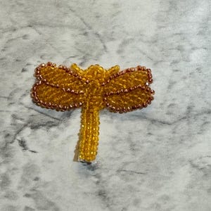 May include: A beaded dragonfly pin in shades of yellow and brown. The wings are a light yellow with brown outlining, and the body is a solid yellow. The pin is made of small beads and is likely handmade.