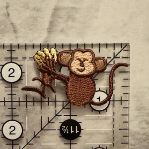 May include: Embroidered patch featuring a brown monkey holding a bunch of yellow bananas. The patch is approximately 1.5 inches wide and 2 inches tall. The monkey has a smiling face and is holding the bananas in its hands. The patch is on a clear ruler.