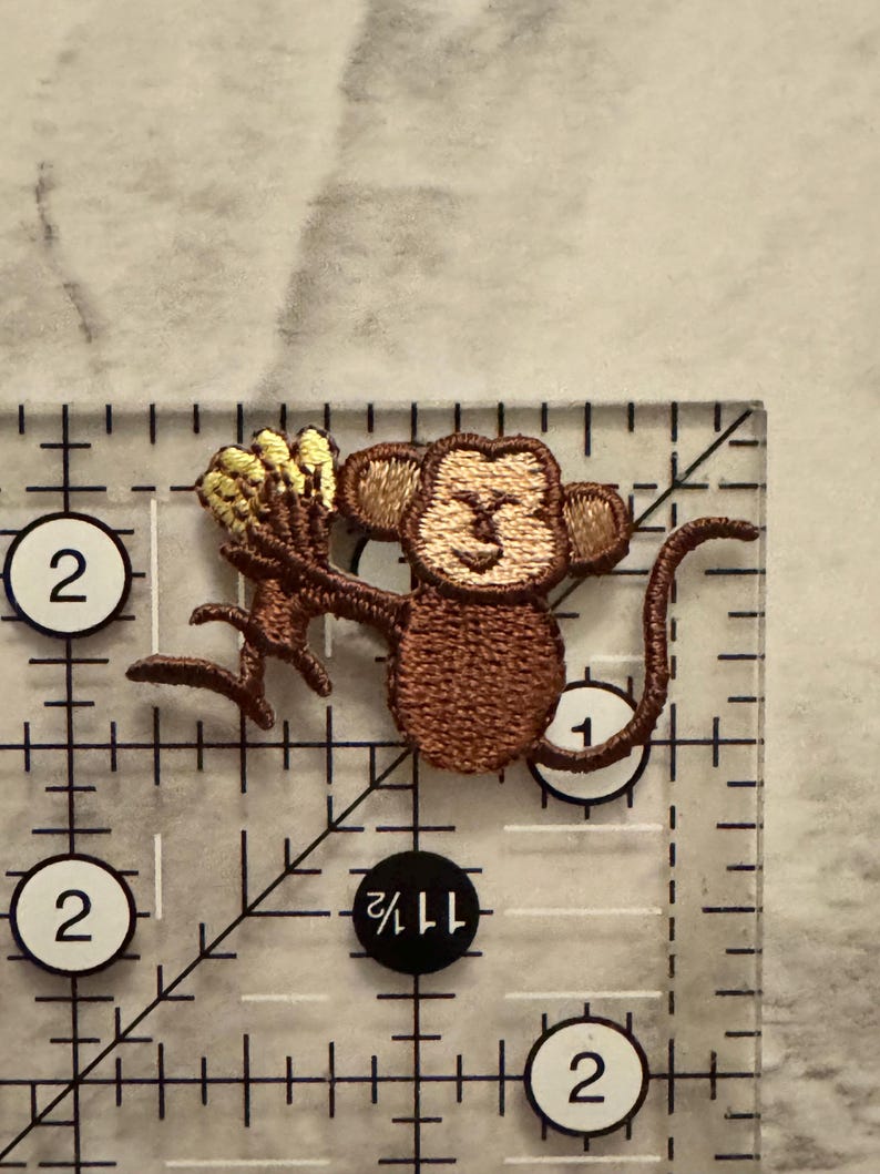 May include: Embroidered patch featuring a brown monkey holding a bunch of yellow bananas. The patch is approximately 1.5 inches wide and 2 inches tall. The monkey has a smiling face and is holding the bananas in its hands. The patch is on a clear ruler.
