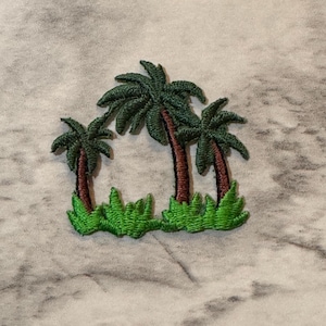 May include: Embroidered patch featuring three palm trees with green fronds and brown trunks, set against a backdrop of green grass. The patch is designed for application on clothing or accessories, evoking a tropical theme.
