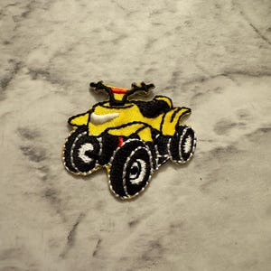 May include: Embroidered patch of a yellow ATV with black and white detailing. The ATV has black wheels, a black seat, and orange handlebars. The patch is likely for clothing or accessories.
