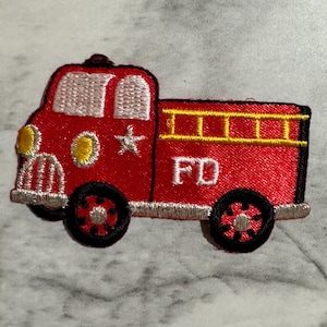 May include: Embroidered patch of a red fire truck with black wheels and yellow ladder detail. The patch features the letters "FD" in white and a star and circle accents. The patch is designed for sewing or ironing onto fabric.