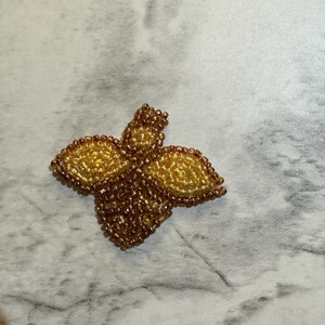 May include: A beaded, four-petal flower pin in shades of gold and brown. The petals are outlined with a darker brown, while the center and edges are a lighter gold. The pin is likely handmade and suitable for clothing or accessories.