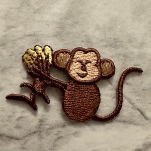 May include: Embroidered patch of a brown monkey holding a bunch of yellow bananas. The monkey has a light brown face and ears, with a curved tail. The patch is likely for clothing or accessories.