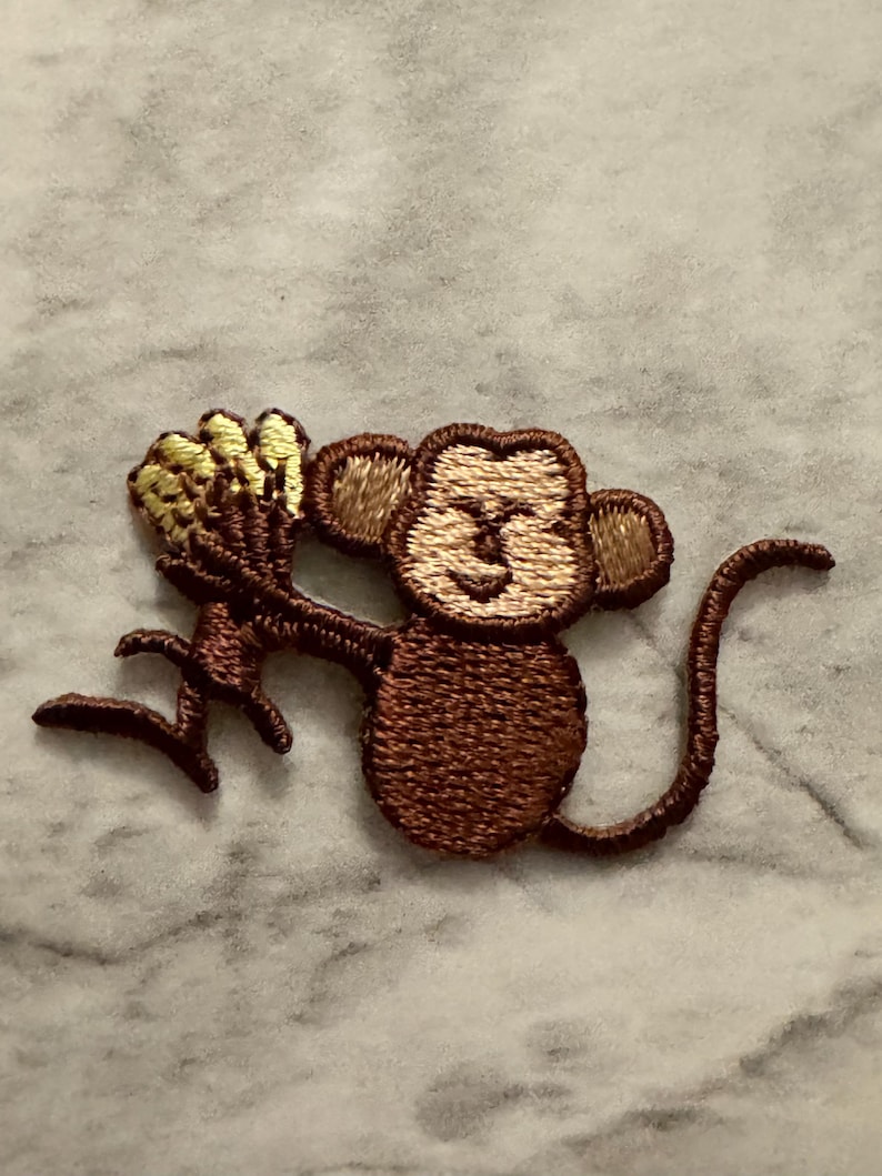 May include: Embroidered patch of a brown monkey holding a bunch of yellow bananas. The monkey has a light brown face and ears, with a curved tail. The patch is likely for clothing or accessories.