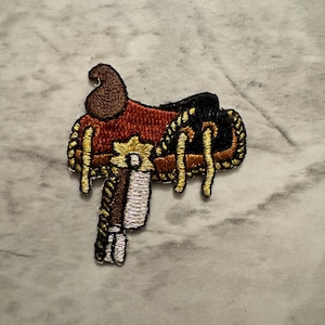 May include: Embroidered patch of a Western saddle. The patch features a brown seat, black accents, and gold-colored decorative stitching. The stirrup is white and silver. The patch is designed to be ironed on or sewn onto fabric.