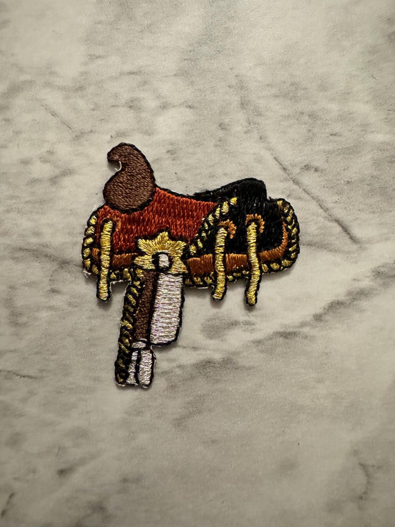 May include: Embroidered patch of a Western saddle. The patch features a brown seat, black accents, and gold-colored decorative stitching. The stirrup is white and silver. The patch is designed to be ironed on or sewn onto fabric.