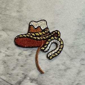 May include: Embroidered patch featuring a cowboy hat and lasso. The hat is brown with a silver and gold trim. The lasso is silver and gold, and the patch is on a gray background. This patch is suitable for clothing or accessories.