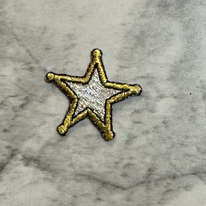 May include: A gold and silver star-shaped patch with a black outline. The star has a textured silver center and gold trim. The patch is likely for decorative purposes, such as on clothing or accessories.