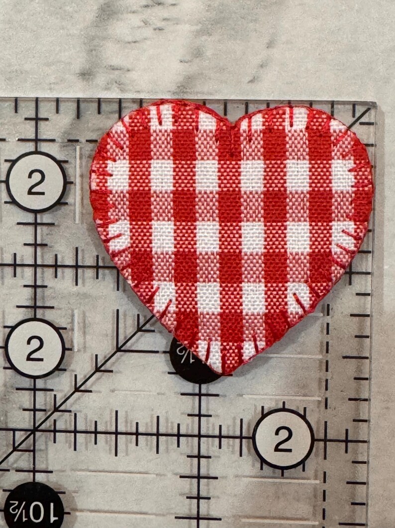May include: A red and white gingham heart-shaped fabric patch. The patch is approximately 2 inches wide and is placed on a clear ruler with black markings and the number 2.