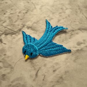 May include: A detailed, embroidered patch of a blue bird in flight. The bird features a bright blue body with darker blue detailing on the wings and tail. The beak is yellow. The patch is designed to be sewn or ironed onto fabric.