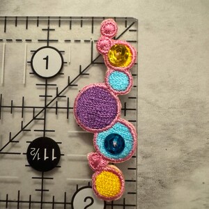 K&ouml;nnte beinhalten: Ein farbenfrohes, gesticktes Applikationsmuster mit einer Reihe von verbundenen Kreisen in Rosa, Lila, Gelb und Blau. Jeder Kreis ist mit Pailletten oder Stickereien verziert. Die Applikation befindet sich auf einem transparenten Lineal mit cm-Markierungen.