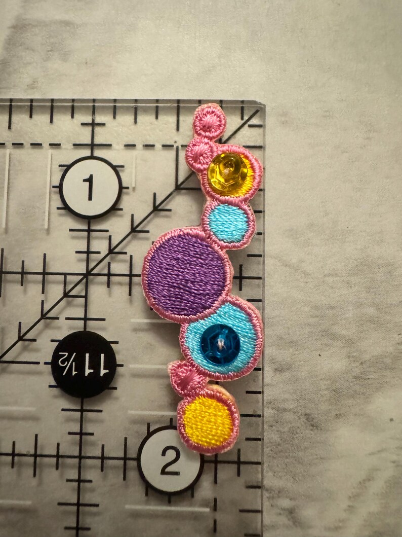 K&ouml;nnte beinhalten: Ein farbenfrohes, gesticktes Applikationsmuster mit einer Reihe von verbundenen Kreisen in Rosa, Lila, Gelb und Blau. Jeder Kreis ist mit Pailletten oder Stickereien verziert. Die Applikation befindet sich auf einem transparenten Lineal mit cm-Markierungen.