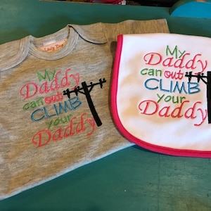 May include: A gray bodysuit and a white bib with pink trim. Both items feature the embroidered text "My Daddy can out climb your Daddy" with a black silhouette of a utility pole and power lines.