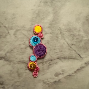 May include: Embroidered patch featuring a vertical arrangement of colorful circles. The circles are in shades of yellow, blue, and purple, with a pink border. Some circles have a shiny gem in the center. This decorative patch is suitable for clothing or accessories.