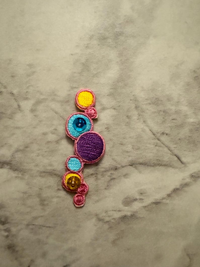 K&ouml;nnte beinhalten: Gestickter Aufn&auml;her mit einer vertikalen Anordnung von bunten Kreisen. Die Kreise sind in Gelb, Blau und Lila gehalten, mit einem rosa Rand. Einige Kreise haben einen gl&auml;nzenden Edelstein in der Mitte. Dieser dekorative Aufn&auml;her eignet sich f&uuml;r Kleidung oder Accessoires.