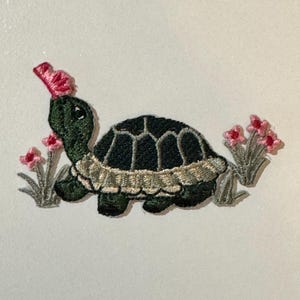 May include: A green and brown embroidered patch of a turtle with a pink flower on its head. The turtle is surrounded by green grass and pink flowers.