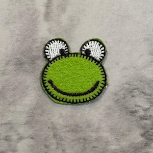 May include: A green frog-shaped patch with a black stitched outline and a smiling mouth. The frog's eyes are white with black pupils and a black stitched outline. The patch is likely for clothing or accessories.