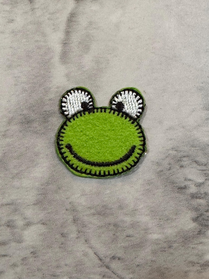 May include: A green frog-shaped patch with a black stitched outline and a smiling mouth. The frog's eyes are white with black pupils and a black stitched outline. The patch is likely for clothing or accessories.