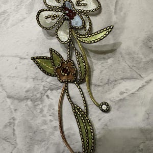 May include: A decorative floral applique featuring a large cream-colored flower with a light blue center and a smaller brown flower below. The design includes green leaves, gold-toned stems, and black embroidered edges with small circular accents.